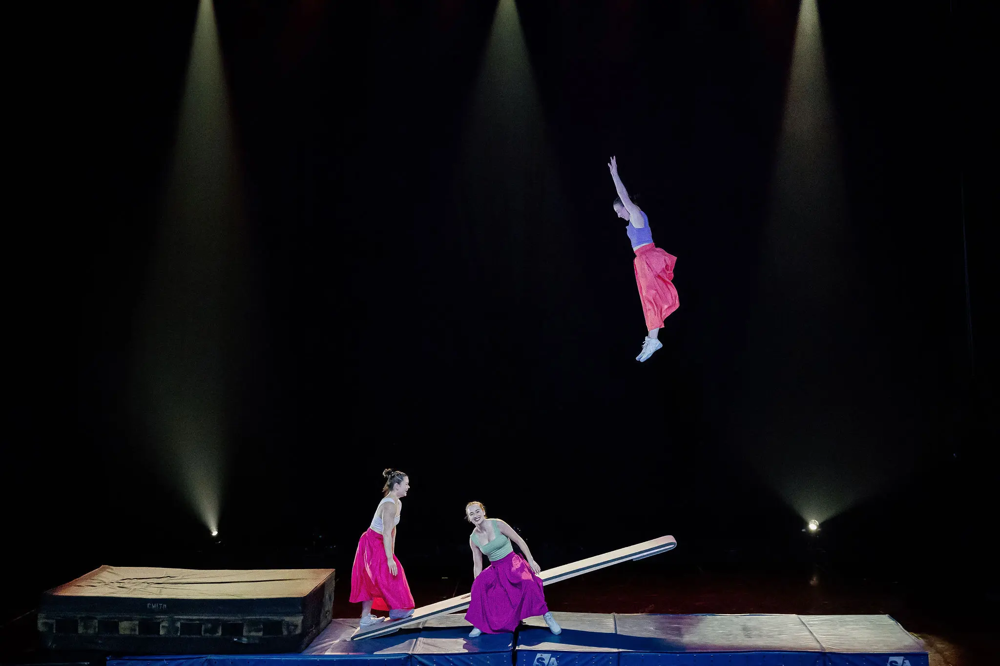 Trois acrobates se produisent sur scène ; Lilyclaire Major-Varallo et Marie-Sophie McKinnon se tiennent sur une bascule tandis qu'une troisième, vêtue d'une jupe rouge, s'envole au milieu d'un saut, sous l'éclairage des projecteurs qui éclairent l'arrière-plan sombre.