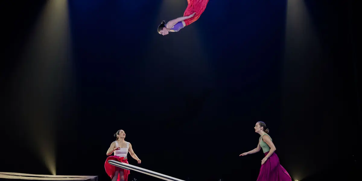 Trois artistes en costumes colorés, dont Lilyclaire Major-Varallo et Marie-Sophie McKinnon, utilisent une bascule pour lancer une acrobate dans les airs sous un éclairage dramatique, tandis que deux autres l'observent et se préparent à la rattraper.