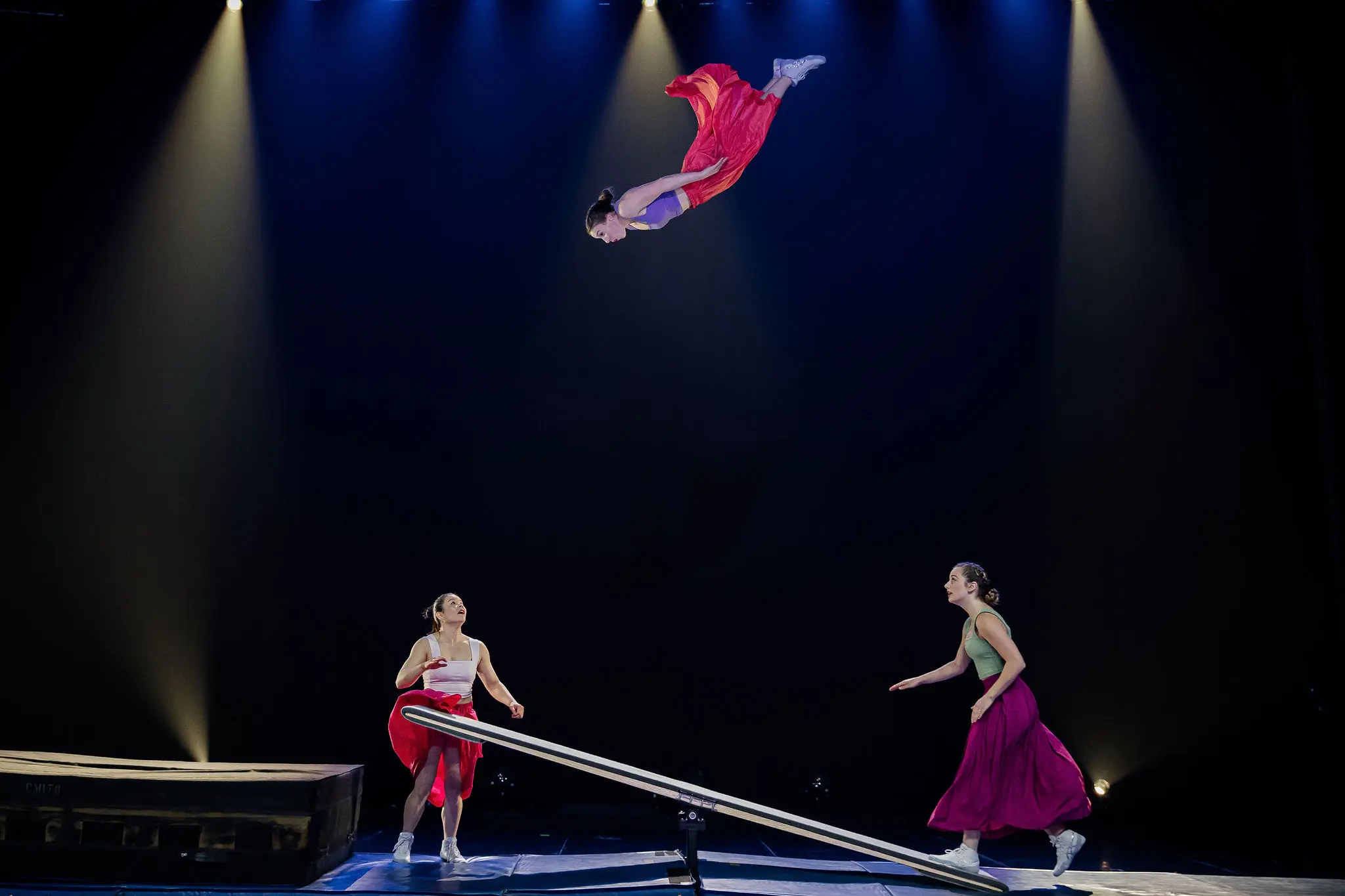 Trois artistes en costumes colorés, dont Lilyclaire Major-Varallo et Marie-Sophie McKinnon, utilisent une bascule pour lancer une acrobate dans les airs sous un éclairage dramatique, tandis que deux autres l'observent et se préparent à la rattraper.