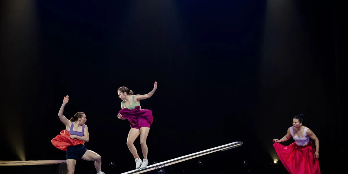 Lilyclaire Major-Varallo et Marie-Sophie McKinnon, vêtues de jupes colorées et de hauts athlétiques, réalisent des prouesses audacieuses sur une planche à bascule. L'une des acrobates s'élève dans les airs tandis que les autres se tiennent en équilibre, l'éclairage dramatique mettant en valeur leurs mouvements habiles.