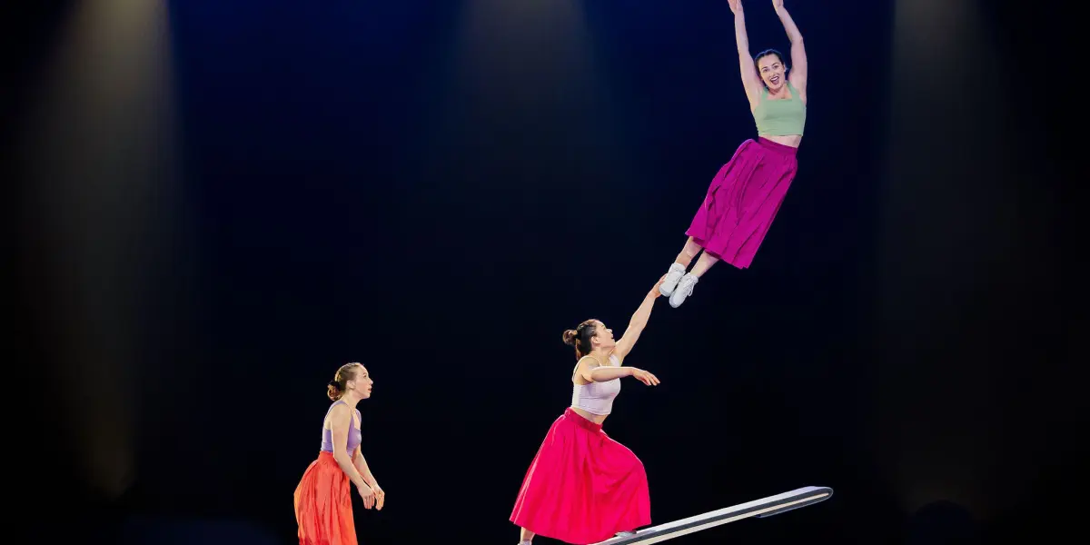 Trois femmes vêtues de jupes colorées se produisent sur un appareil ressemblant à une balançoire sous les lumières de la scène ; Lilyclaire Major-Varallo est lancée haut dans les airs, les bras levés, tandis que Marie-Sophie McKinnon et une troisième la soutiennent.