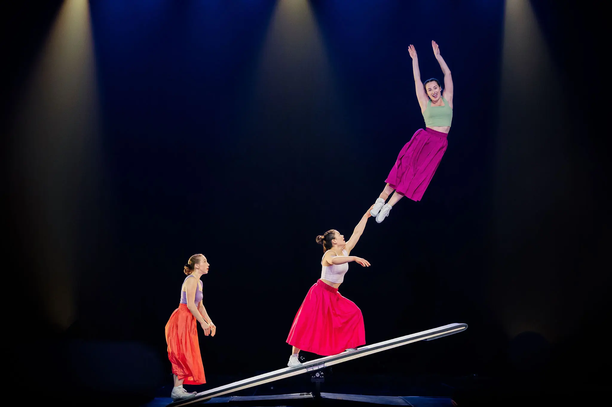 Trois femmes vêtues de jupes colorées se produisent sur un appareil ressemblant à une balançoire sous les lumières de la scène ; Lilyclaire Major-Varallo est lancée haut dans les airs, les bras levés, tandis que Marie-Sophie McKinnon et une troisième la soutiennent.