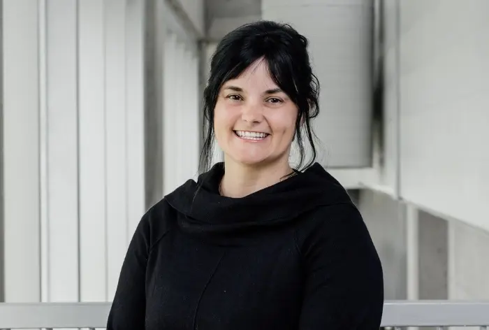 Geneviève Dupéré, une femme aux cheveux noirs attachés, vêtue d'un haut noir, sourit alors qu'elle se tient à l'intérieur sur un fond de murs blancs et gris.