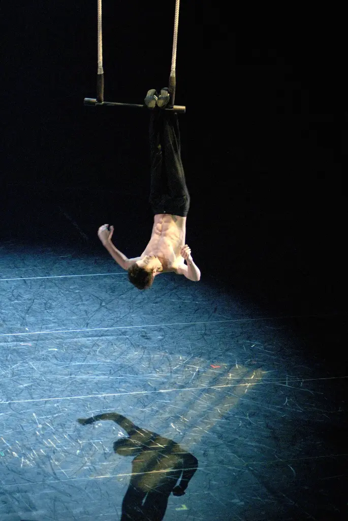 Un acrobate torse nu, Bret Pfister, est suspendu par les jambes à un trapèze sous un projecteur, projetant une ombre sur le sol sombre de la scène.