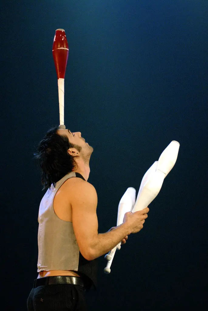 Un artiste de cirque, Gonzalo Coloma Rospigliosi, tient en équilibre une massue de jonglage rouge sur son front et deux massues blanches dans ses mains sur un fond bleu foncé.