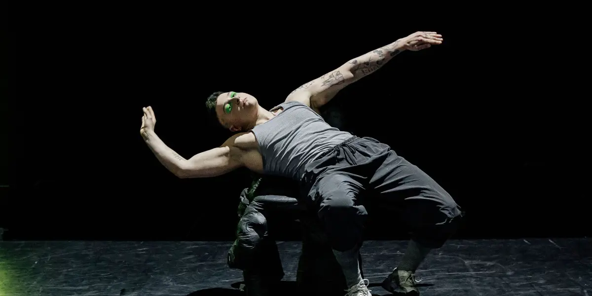 Nin Khelifa, une danseuse vêtue d'un débardeur gris et d'un pantalon sombre, se penche dramatiquement en arrière sur une chaise sur une scène faiblement éclairée, les bras étendus au milieu de la représentation.