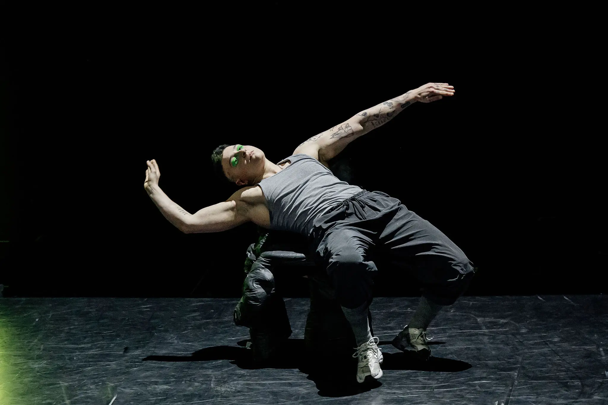 Nin Khelifa, une danseuse vêtue d'un débardeur gris et d'un pantalon sombre, se penche dramatiquement en arrière sur une chaise sur une scène faiblement éclairée, les bras étendus au milieu de la représentation.