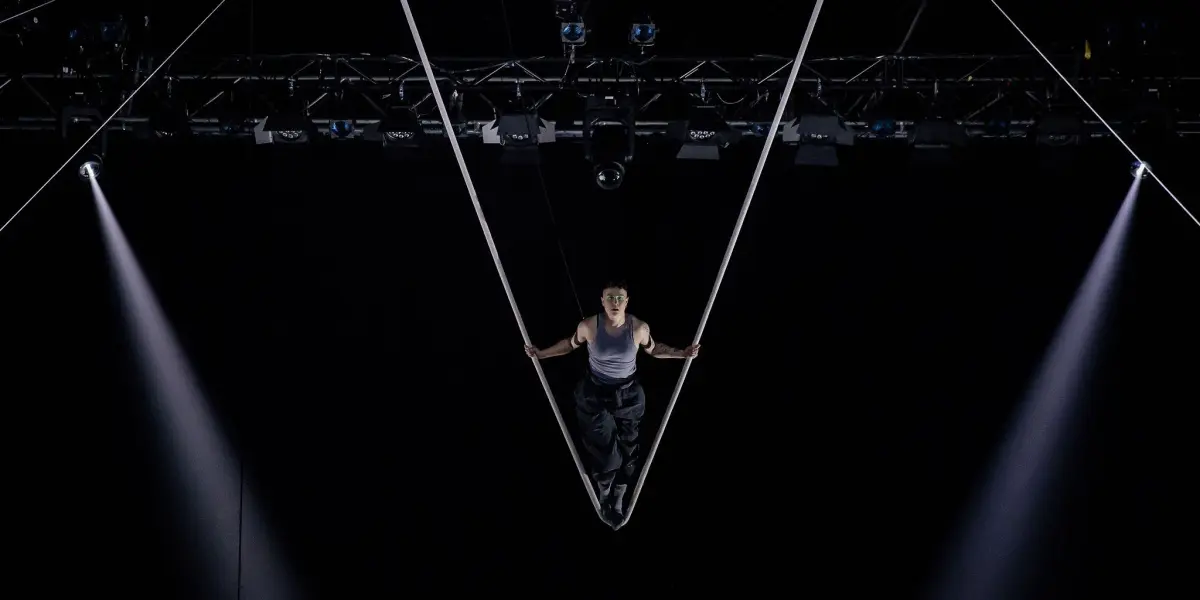 Nin Khelifa se produit sur des sangles aériennes, suspendues en forme de V au-dessus d'une scène sombre, avec des projecteurs qui brillent de part et d'autre.