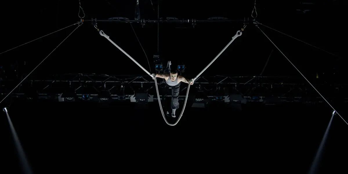 L'acrobate de cirque Nin Khelifa est suspendu dans les airs, se produisant sur des anneaux de gymnastique, les bras tendus en forme de V, sur un fond sombre où l'on aperçoit des lumières de scène et des câbles.