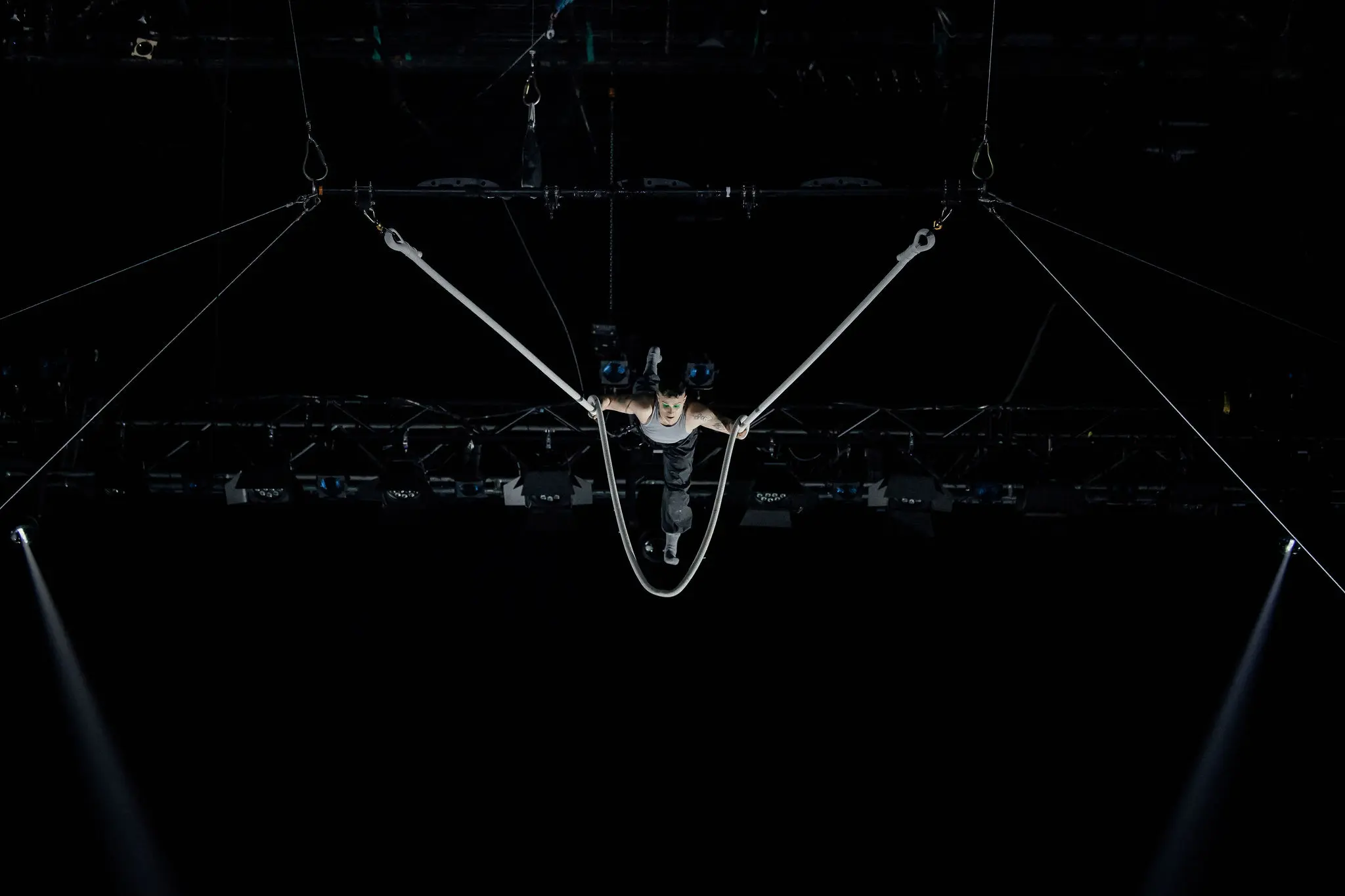 L'acrobate de cirque Nin Khelifa est suspendu dans les airs, se produisant sur des anneaux de gymnastique, les bras tendus en forme de V, sur un fond sombre où l'on aperçoit des lumières de scène et des câbles.