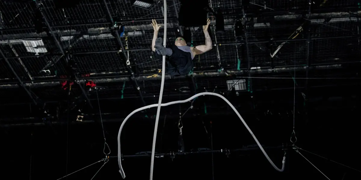 Nin Khelifa exécute un numéro d'acrobatie aérienne en hauteur, suspendu par de longues cordes, avec une scène sombre et un équipement d'éclairage visible au-dessus de sa tête.