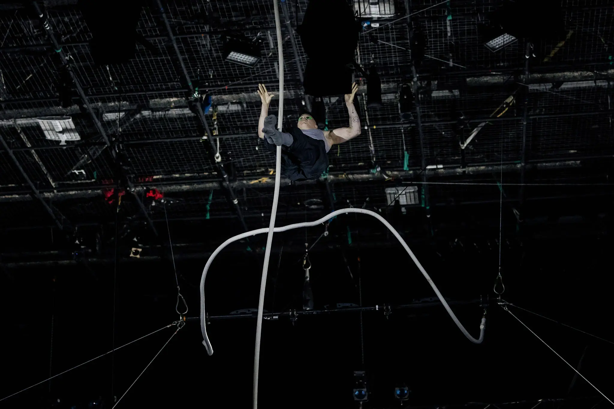 Nin Khelifa exécute un numéro d'acrobatie aérienne en hauteur, suspendu par de longues cordes, avec une scène sombre et un équipement d'éclairage visible au-dessus de sa tête.