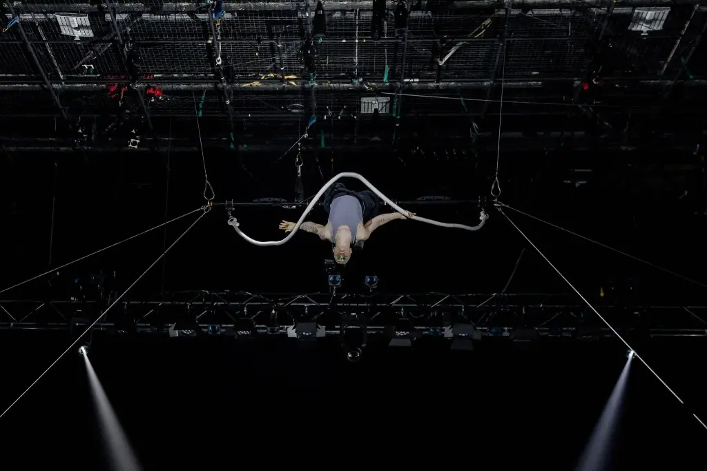 Nin Khelifa exécute un numéro d'acrobatie aérienne, suspendu au plafond par une corde en boucle, avec des lumières de scène et des gréements visibles au-dessus d'eux sur un fond sombre.