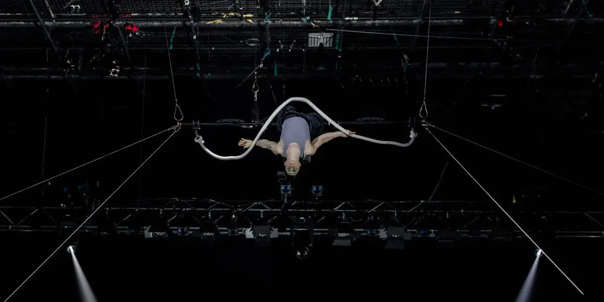 Nin Khelifa exécute un numéro d'acrobatie aérienne, suspendu au plafond par une corde en boucle, avec des lumières de scène et des gréements visibles au-dessus d'eux sur un fond sombre.