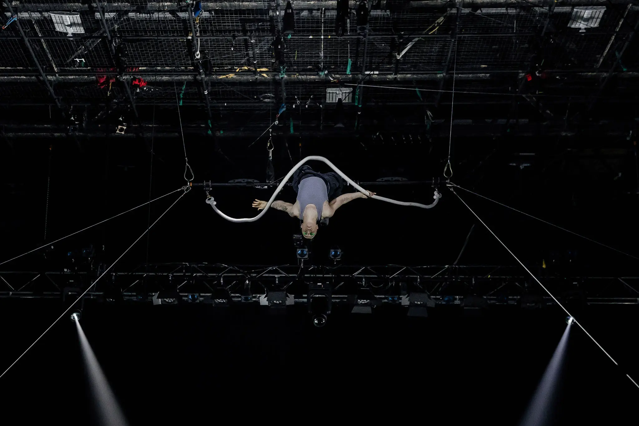 Nin Khelifa exécute un numéro d'acrobatie aérienne, suspendu au plafond par une corde en boucle, avec des lumières de scène et des gréements visibles au-dessus d'eux sur un fond sombre.