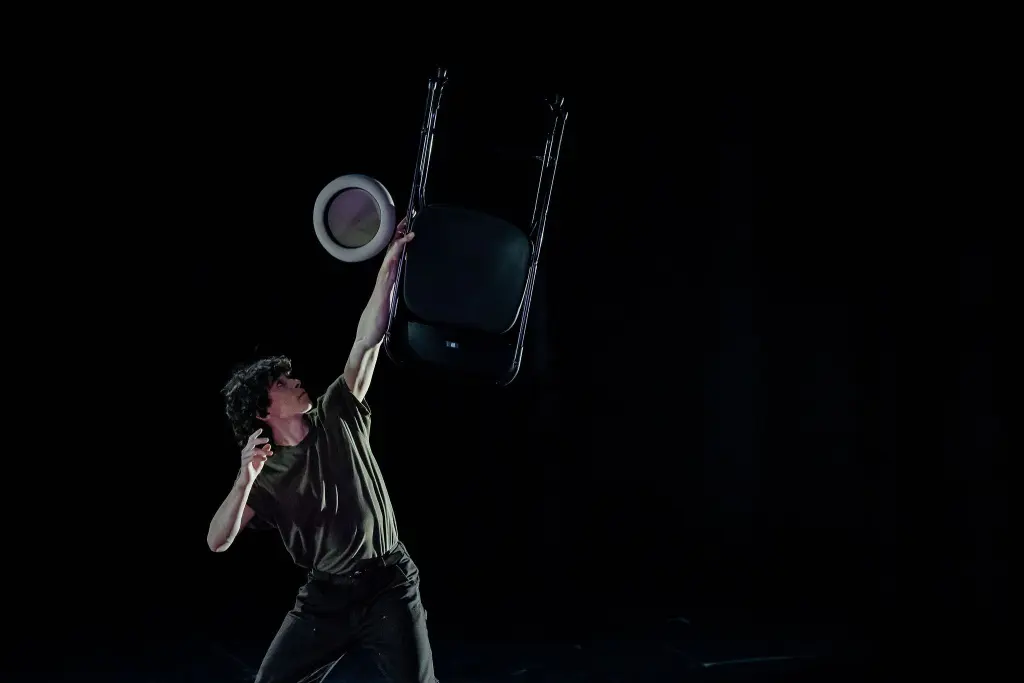 William Borges, vêtu d'un t-shirt sombre, tient une chaise pliante en équilibre sur une main tout en tendant l'autre vers un chapeau blanc sur un fond sombre et dramatique.