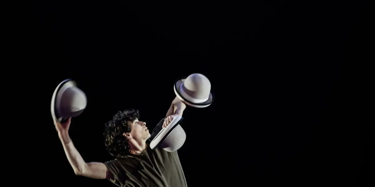 Un artiste vêtu de vêtements sombres, dans le style de William Borges, balance de façon spectaculaire deux chapeaux, un dans chaque main, tout en se tenant à côté d'une chaise noire sur une scène faiblement éclairée avec un arrière-plan sombre.