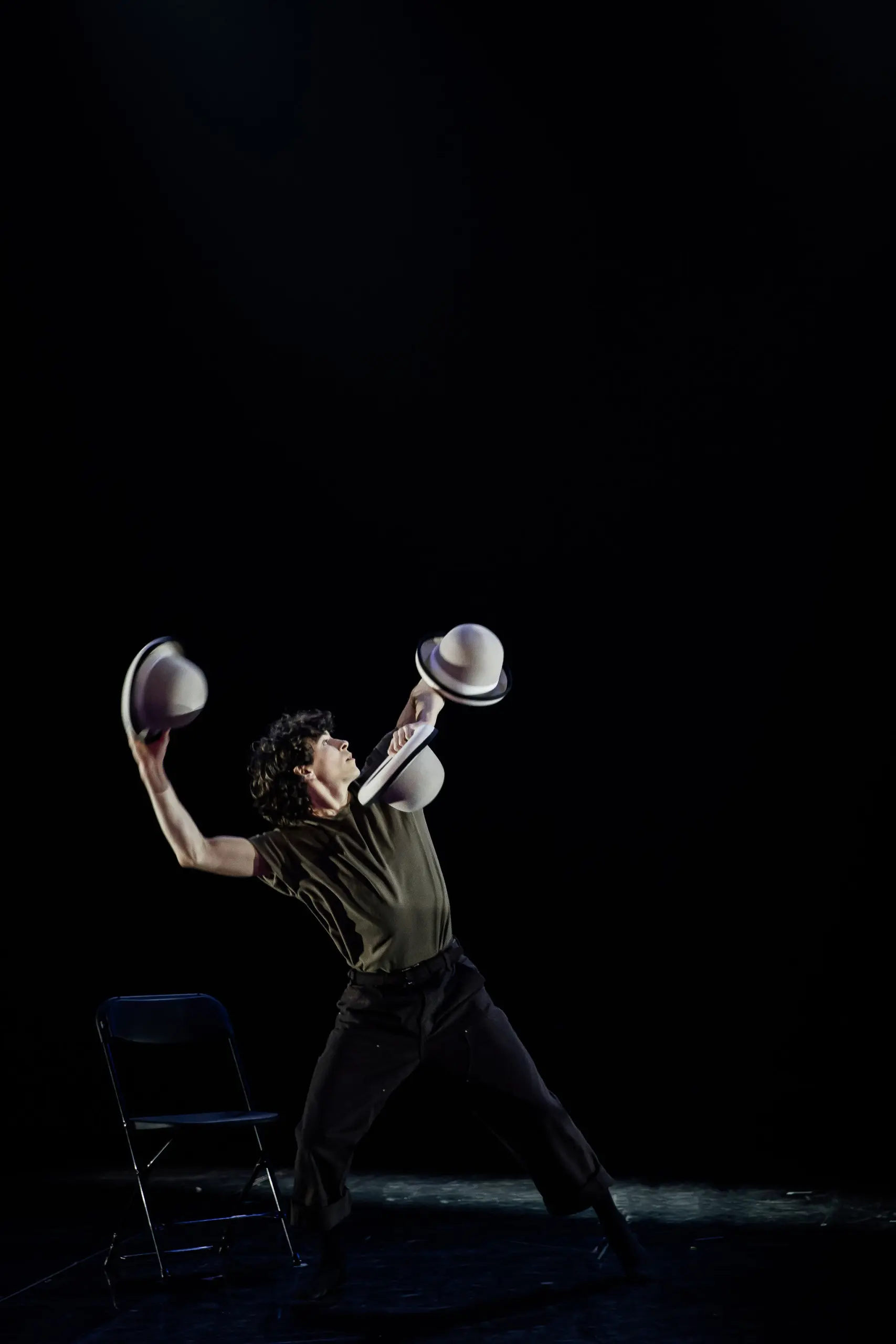 Un artiste vêtu de vêtements sombres, dans le style de William Borges, balance de façon spectaculaire deux chapeaux, un dans chaque main, tout en se tenant à côté d'une chaise noire sur une scène faiblement éclairée avec un arrière-plan sombre.