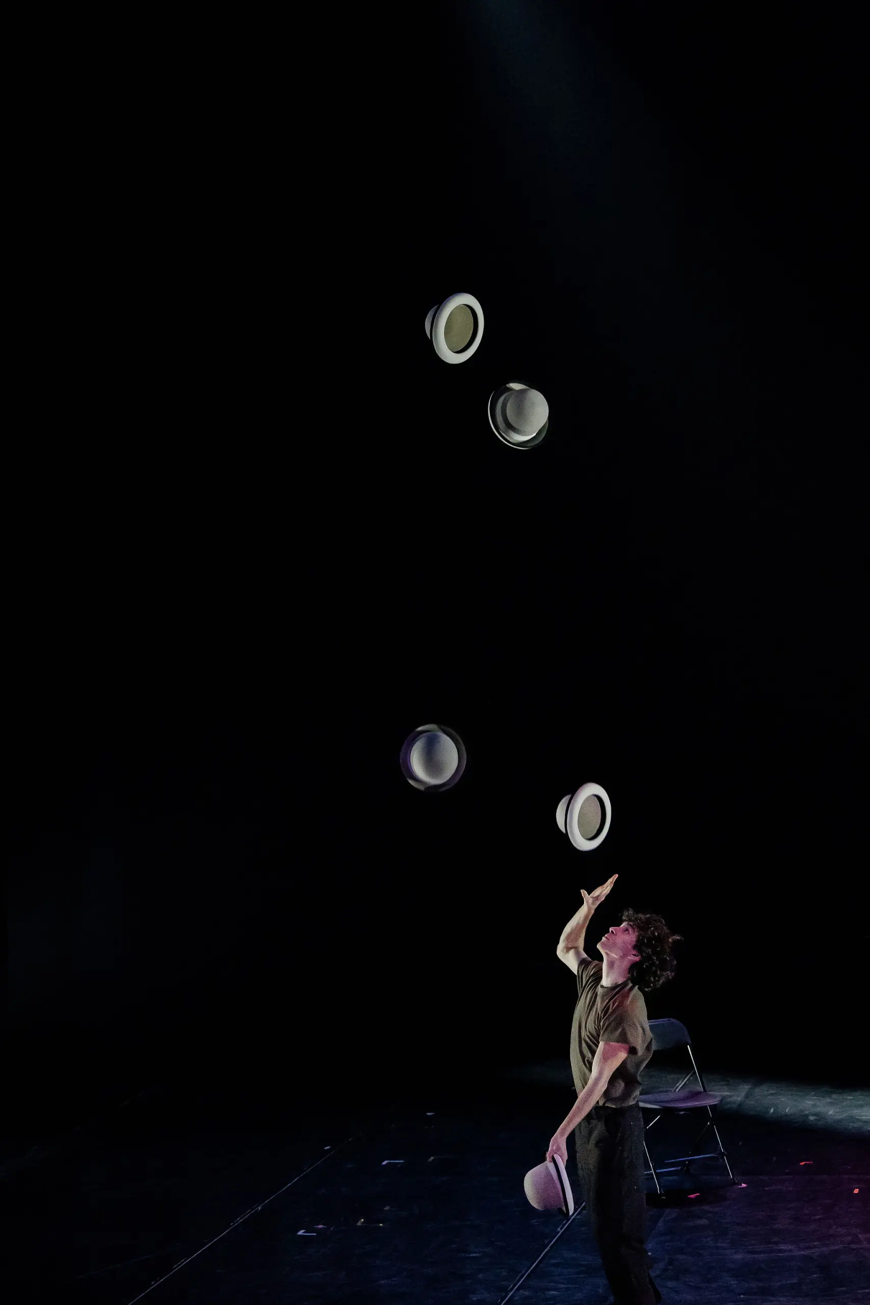William Borges, un artiste sur une scène faiblement éclairée, jongle habilement avec quatre chapeaux blancs dans les airs, avec une chaise vide en arrière-plan et un éclairage dramatique soulignant l'action.