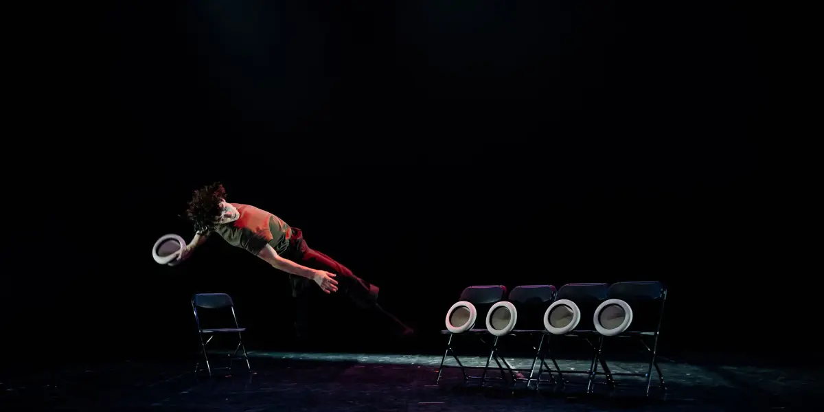 Un interprète saute latéralement en l'air, un chapeau à la main, devant une rangée de cinq chaises noires - chacune sur laquelle est posé un chapeau blanc - sur une scène sombre et dramatique, rappelant le style captivant de William Borges.