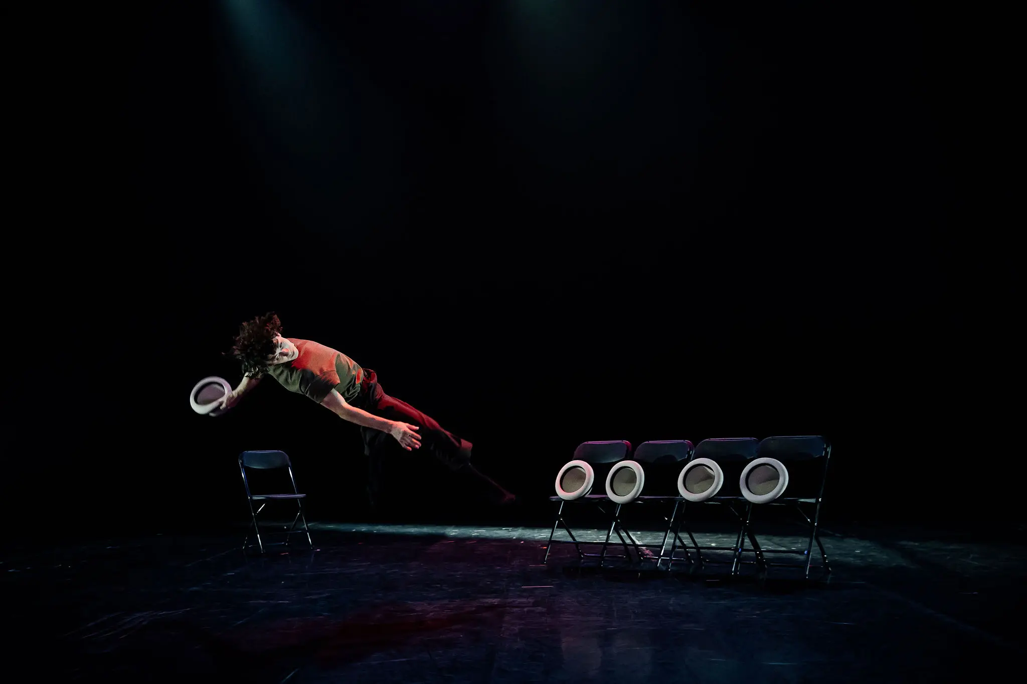 Un interprète saute latéralement en l'air, un chapeau à la main, devant une rangée de cinq chaises noires - chacune sur laquelle est posé un chapeau blanc - sur une scène sombre et dramatique, rappelant le style captivant de William Borges.