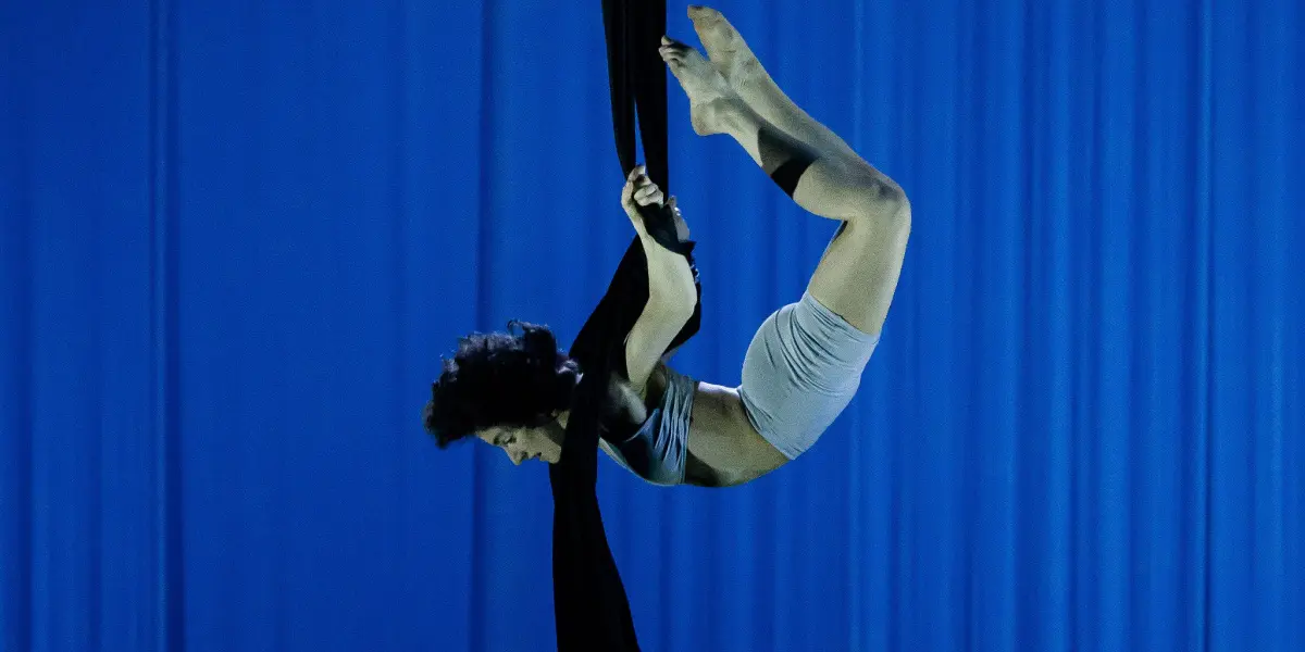 Une voltigeuse de cirque, Sara Sabina Arizmendi Fernandez, en tenue de sport grise, exécute une pose acrobatique, suspendue à l'envers sur des soies noires, sur un fond de scène éclairé en bleu.
