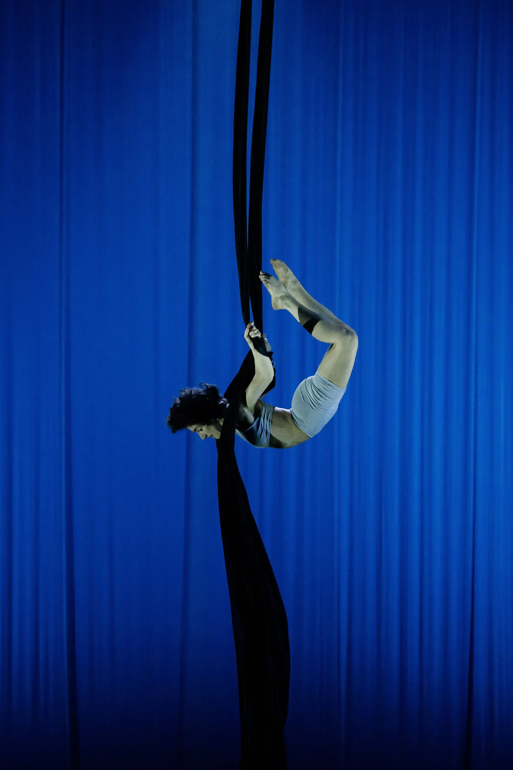 Une voltigeuse de cirque, Sara Sabina Arizmendi Fernandez, en tenue de sport grise, exécute une pose acrobatique, suspendue à l'envers sur des soies noires, sur un fond de scène éclairé en bleu.