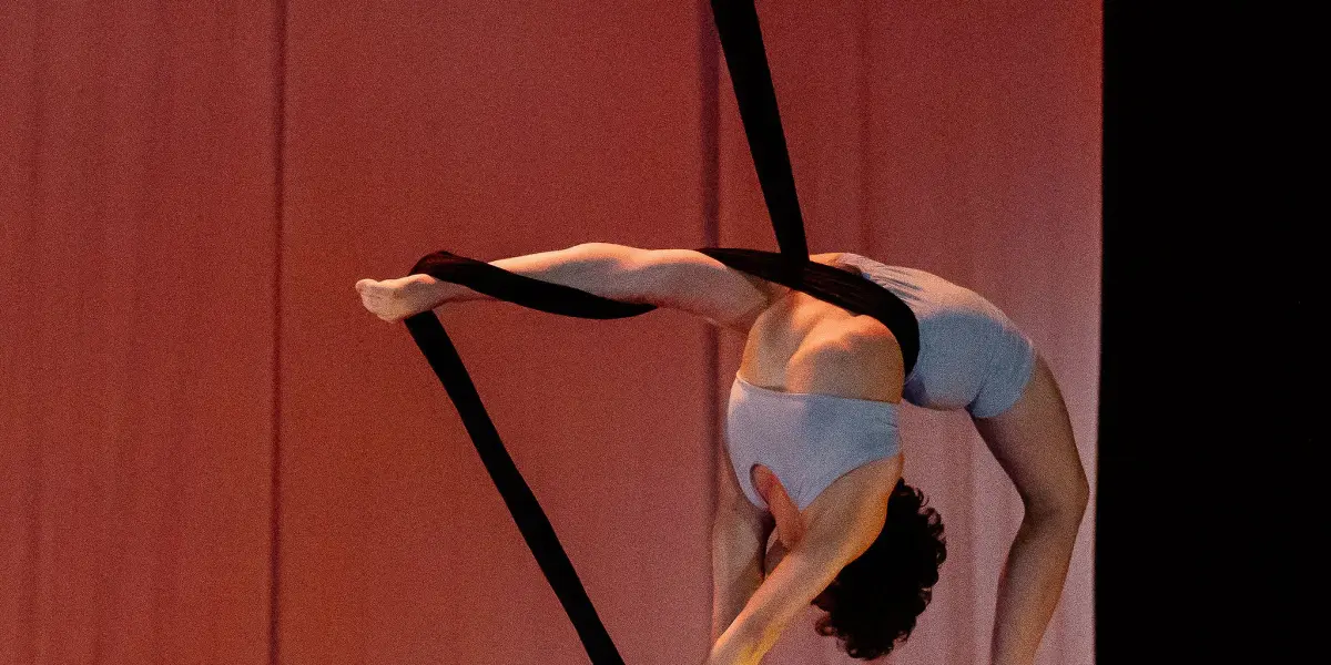 L'artiste aérienne Sara Sabina Arizmendi Fernandez, vêtue d'une tenue blanche, est suspendue la tête en bas, se balançant gracieusement sur des soies aériennes noires, sur un fond de scène rose et orange légèrement éclairé.