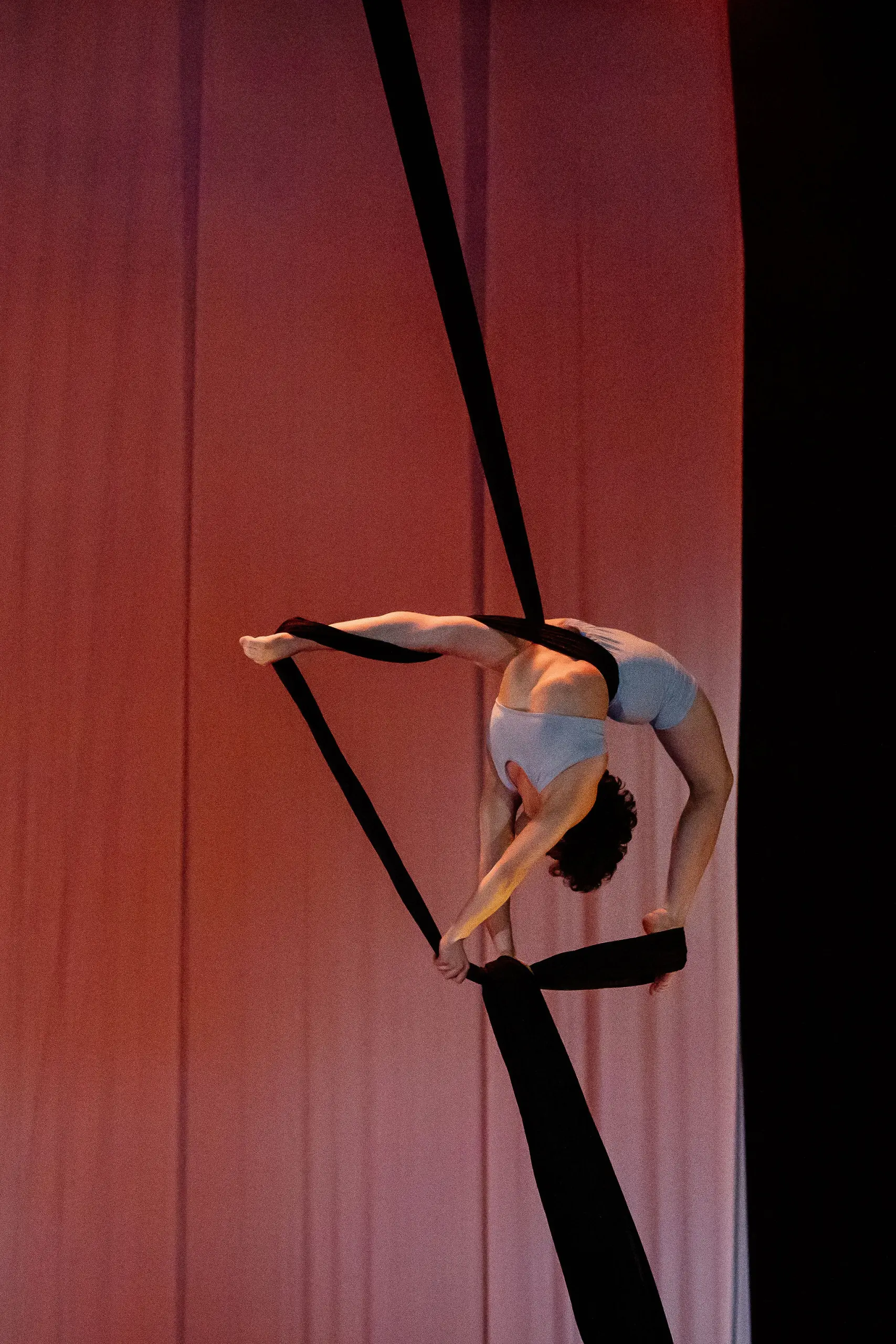 L'artiste aérienne Sara Sabina Arizmendi Fernandez, vêtue d'une tenue blanche, est suspendue la tête en bas, se balançant gracieusement sur des soies aériennes noires, sur un fond de scène rose et orange légèrement éclairé.