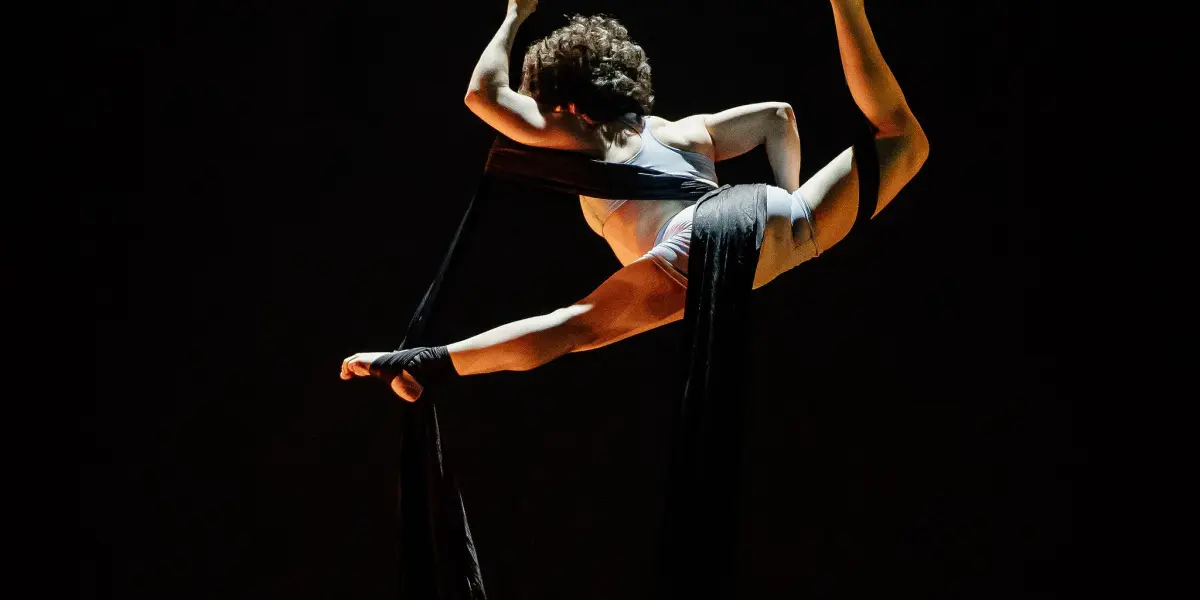L'artiste aérienne Sara Sabina Arizmendi Fernandez, vêtue d'une tenue blanche, est gracieusement suspendue dans les airs, enveloppée dans de longs rubans de soie noire, sur un fond sombre, avec un éclairage dramatique mettant en valeur sa pose.