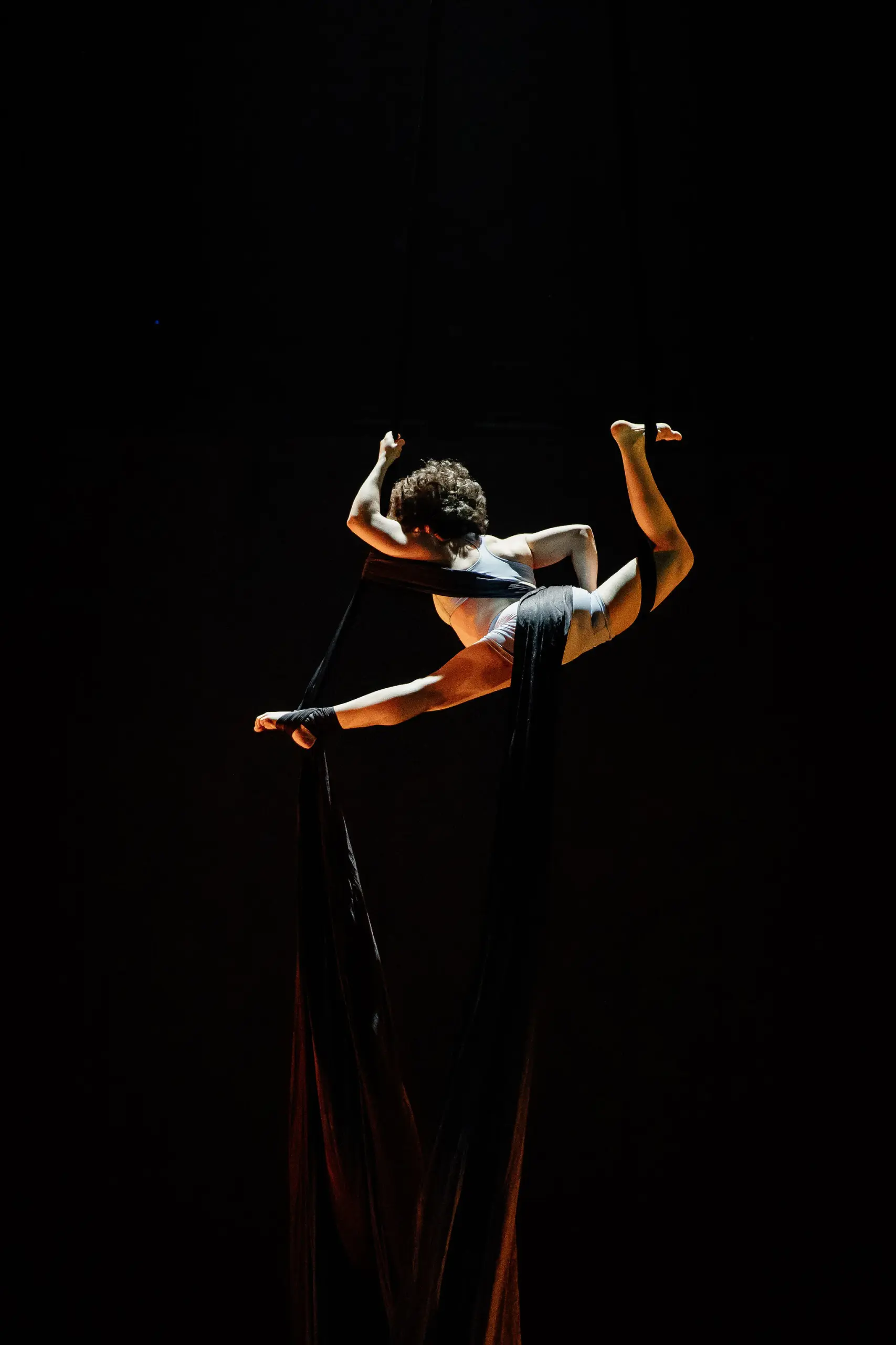 L'artiste aérienne Sara Sabina Arizmendi Fernandez, vêtue d'une tenue blanche, est gracieusement suspendue dans les airs, enveloppée dans de longs rubans de soie noire, sur un fond sombre, avec un éclairage dramatique mettant en valeur sa pose.