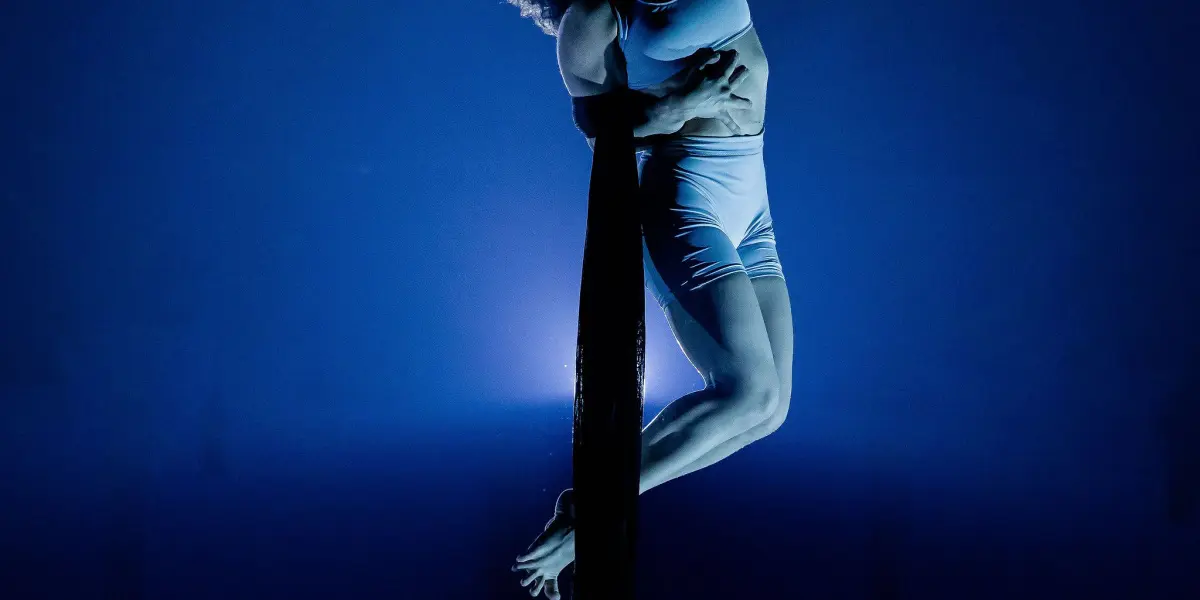 L'acrobate aérienne Sara Sabina Arizmendi Fernandez se produit sur un tissu suspendu, éclairé par une lumière de scène bleue spectaculaire, le corps fléchi et un bras serrant fermement le tissu.