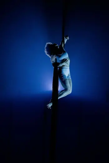 L'acrobate aérienne Sara Sabina Arizmendi Fernandez se produit sur un tissu suspendu, éclairé par une lumière de scène bleue spectaculaire, le corps fléchi et un bras serrant fermement le tissu.