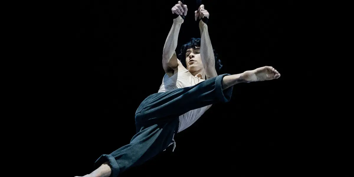 William Borges exécute un numéro aérien, saisissant des sangles à deux mains tout en étant suspendu dans les airs, les jambes étendues en fente sur un fond sombre.