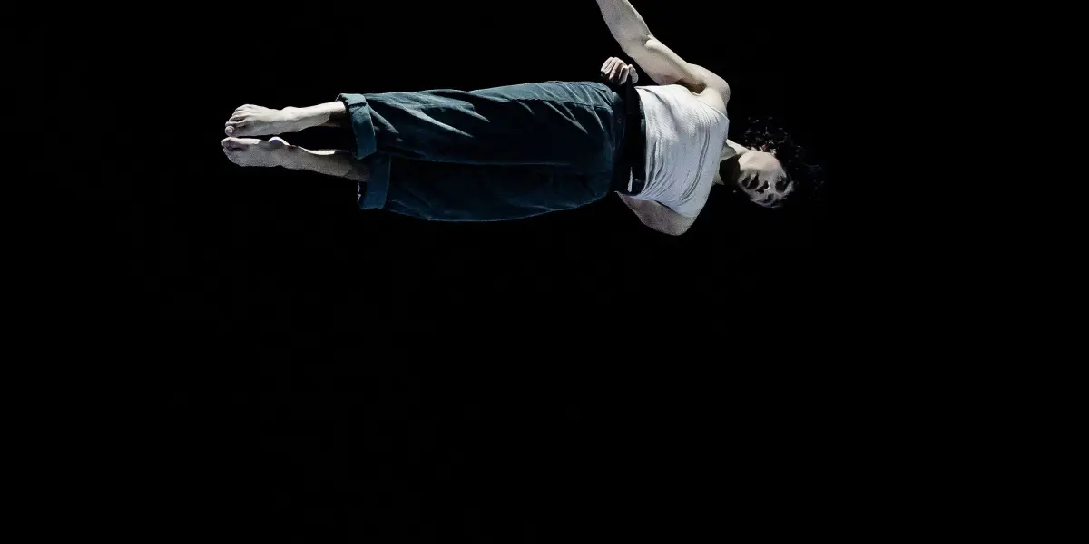 Une personne vêtue d'un pantalon sombre et d'un haut blanc est suspendue horizontalement dans les airs sur un fond noir, se tenant d'une main à une fine corde - une image qui rappelle le style artistique saisissant de William Borges.