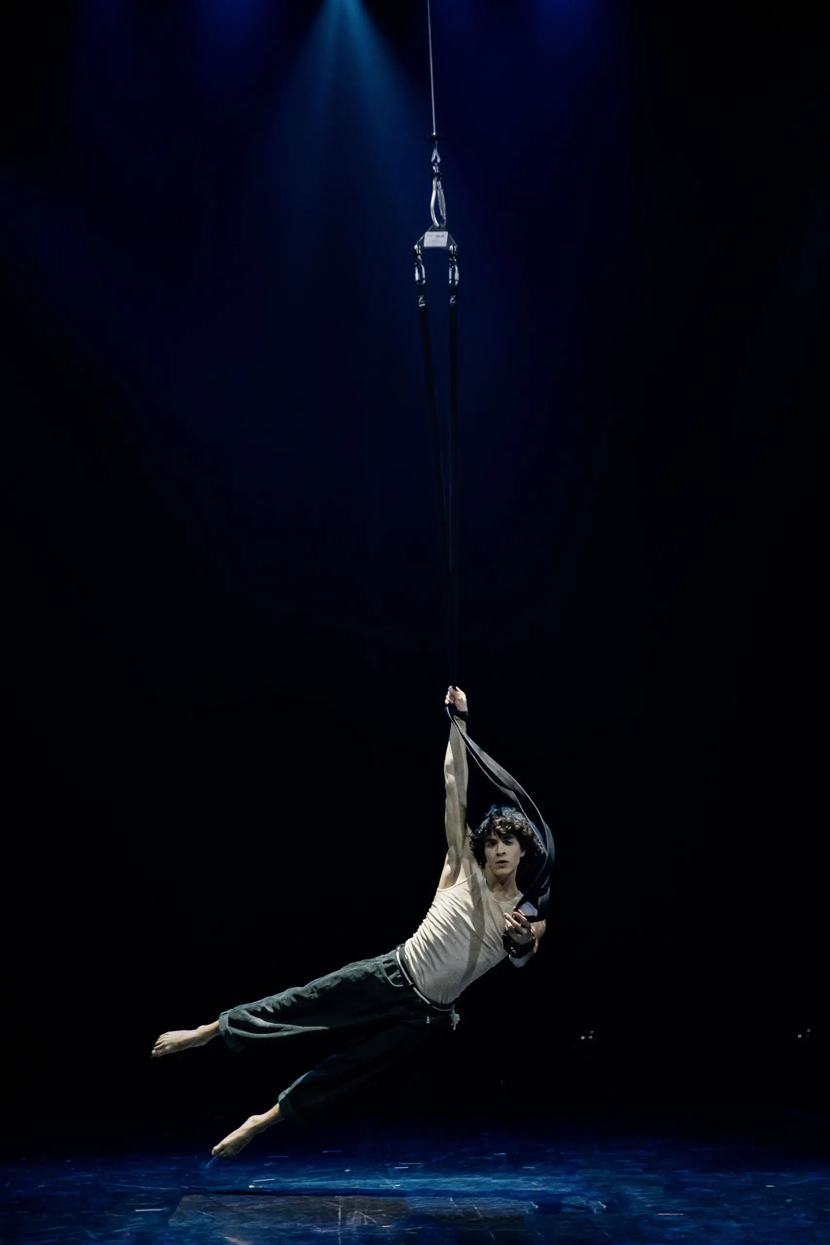 William Borges réalise un numéro d'acrobatie aérienne, suspendu à des sangles de tissu noir, sous un éclairage dramatique et sur une scène sombre.