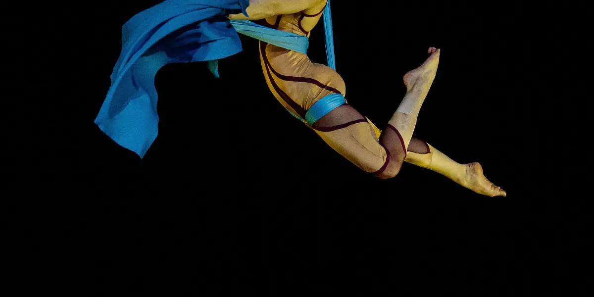 L'artiste aérien Kayley Borckardt Hennessy est suspendu dans les airs dans une pose gracieuse, s'accrochant à un tissu bleu sur fond noir, vêtu d'un costume doré.