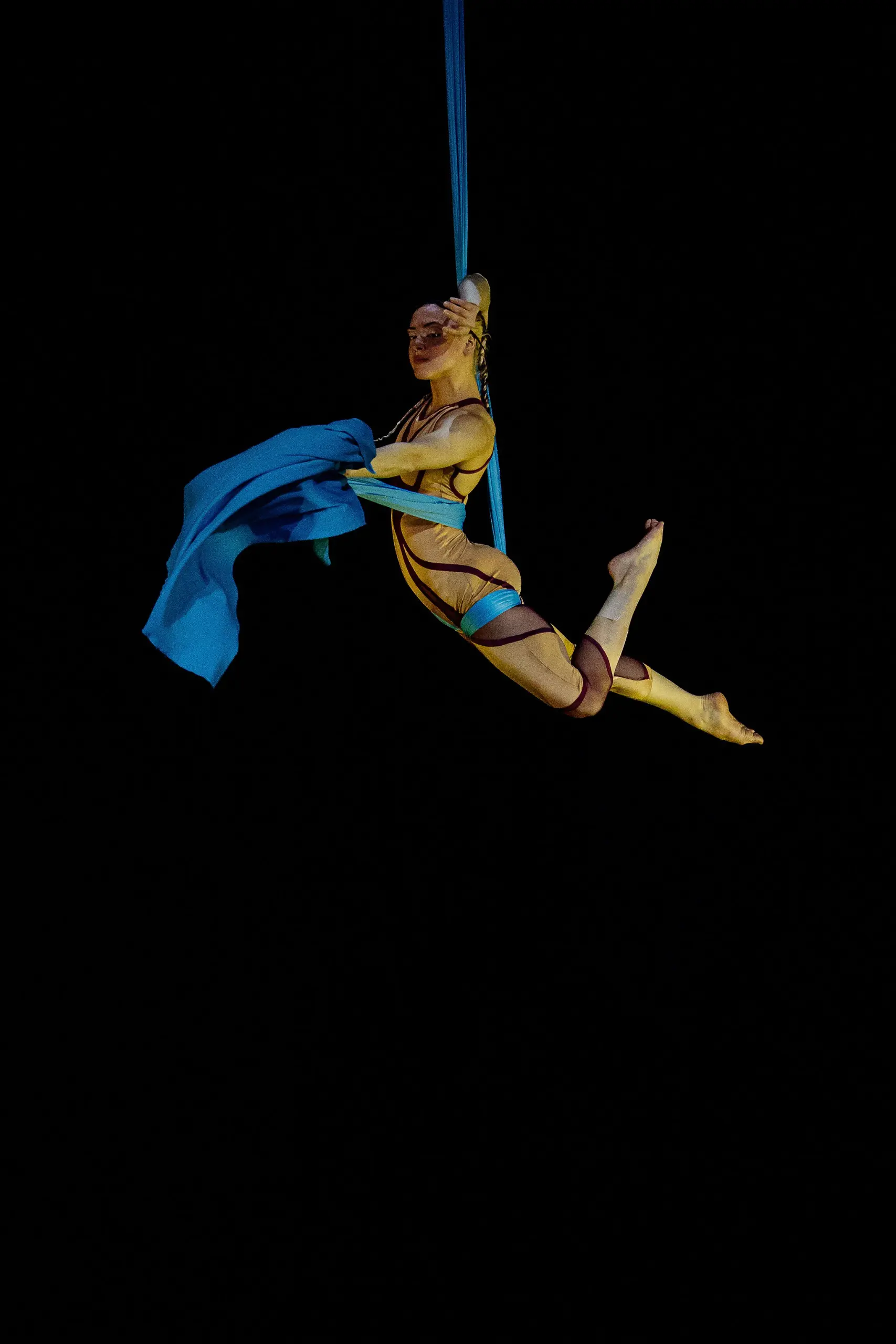 L'artiste aérien Kayley Borckardt Hennessy est suspendu dans les airs dans une pose gracieuse, s'accrochant à un tissu bleu sur fond noir, vêtu d'un costume doré.