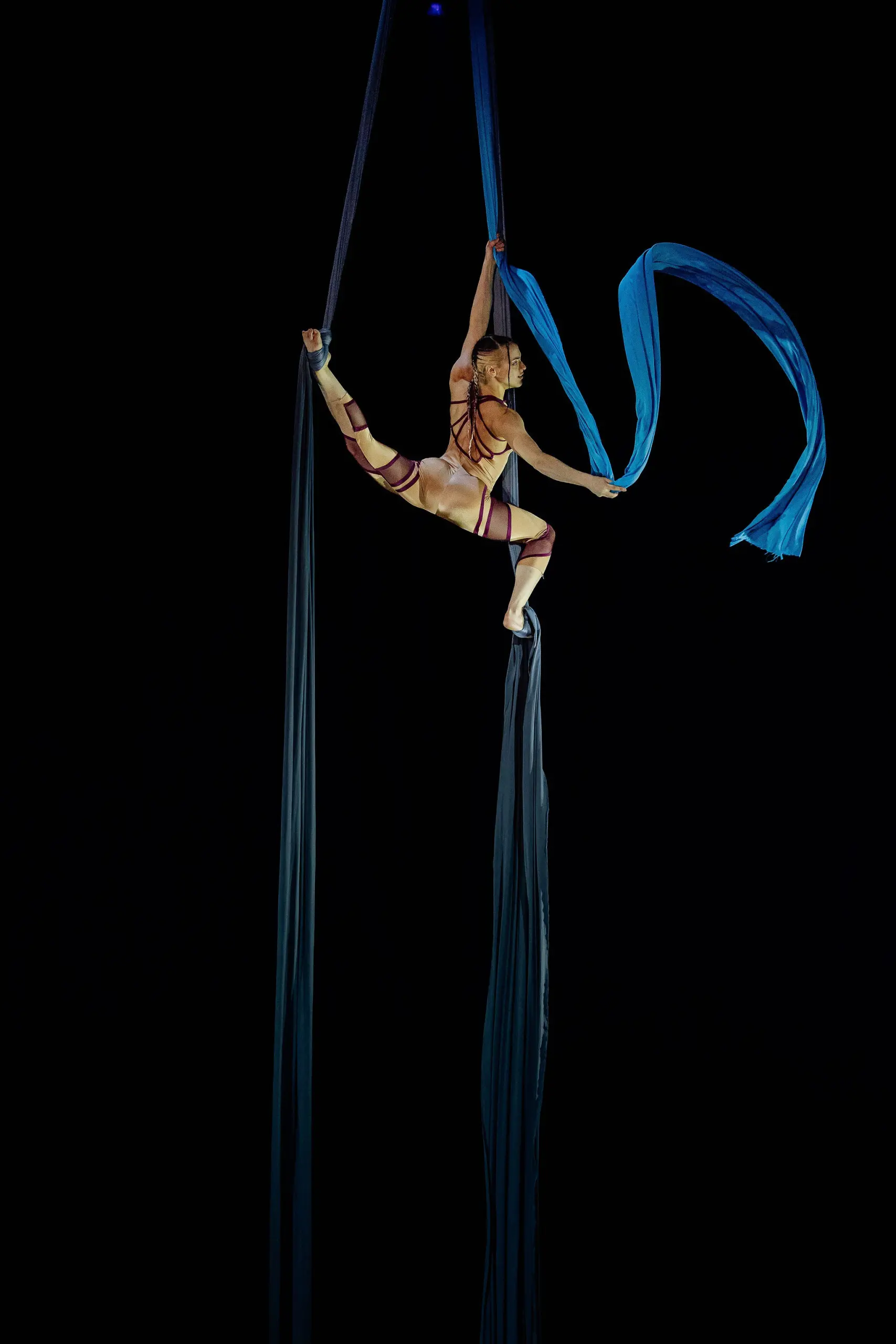 L'artiste aérienne Kayley Borckardt Hennessy est suspendue en l'air sur des soies bleues, prenant une pose fendue avec un bras et une jambe étendus, sur un fond sombre.