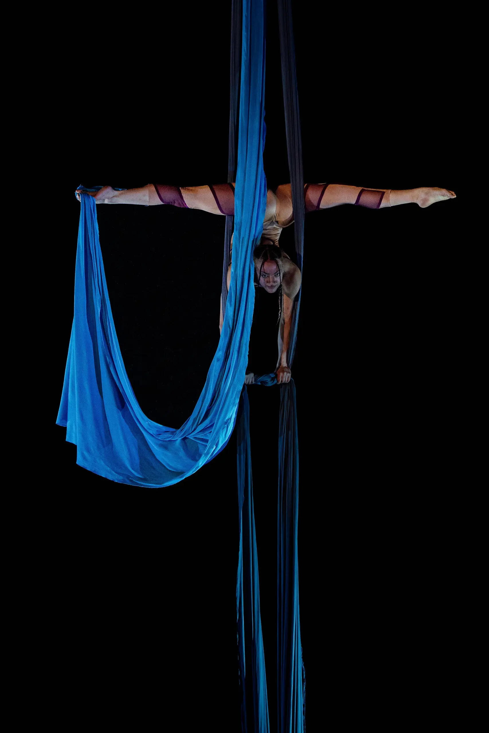 L'artiste aérienne Kayley Borckardt Hennessy exécute un saut en suspension à l'envers sur des soies bleues, en tenant le tissu à deux mains sur un fond noir.