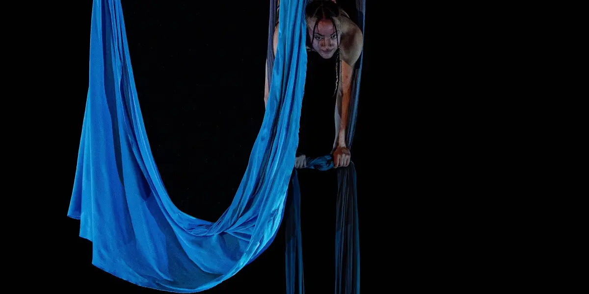 L'acrobate Kayley Borckardt Hennessy exécute un grand écart en se suspendant à l'envers sur un tissu de soie bleu sur fond noir, mettant en valeur une force et une souplesse incroyables.