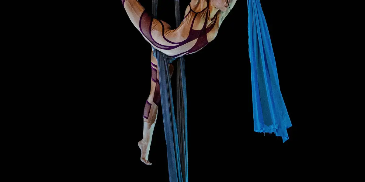 L'artiste aérienne Kayley Borckardt Hennessy, vêtue d'une combinaison à motifs, exécute une pose acrobatique sur des soies bleues, suspendue en l'air sur un fond noir.