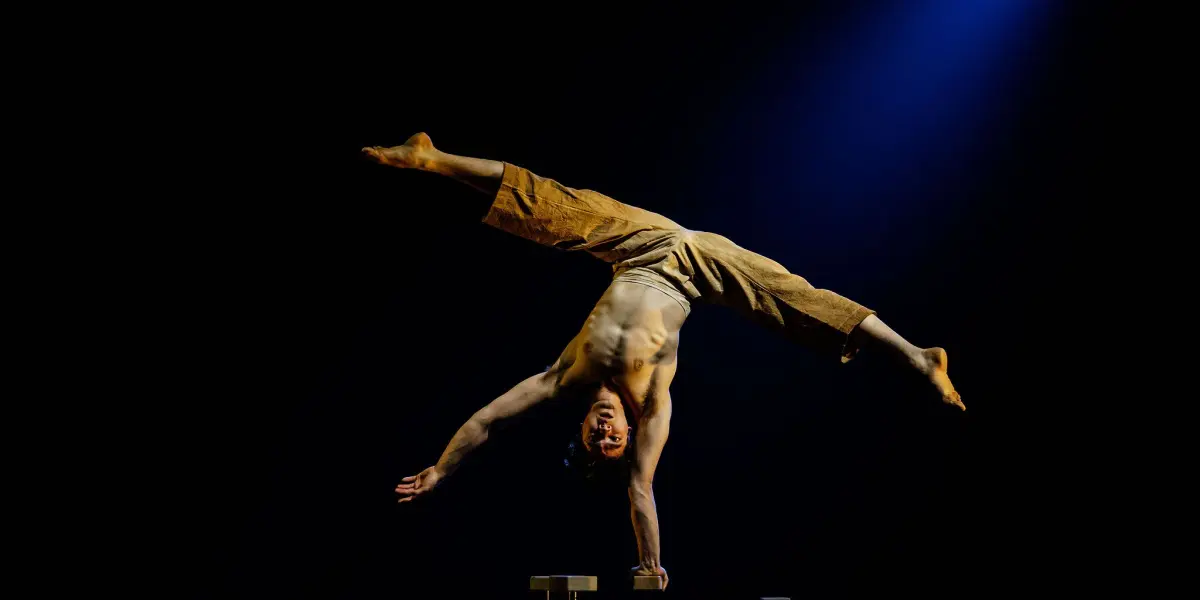 Maël Trân, acrobate torse nu en pantalon marron, effectue un équilibre à une main sur une plate-forme surélevée sous un éclairage de scène spectaculaire, mis en valeur par un projecteur bleu en arrière-plan.