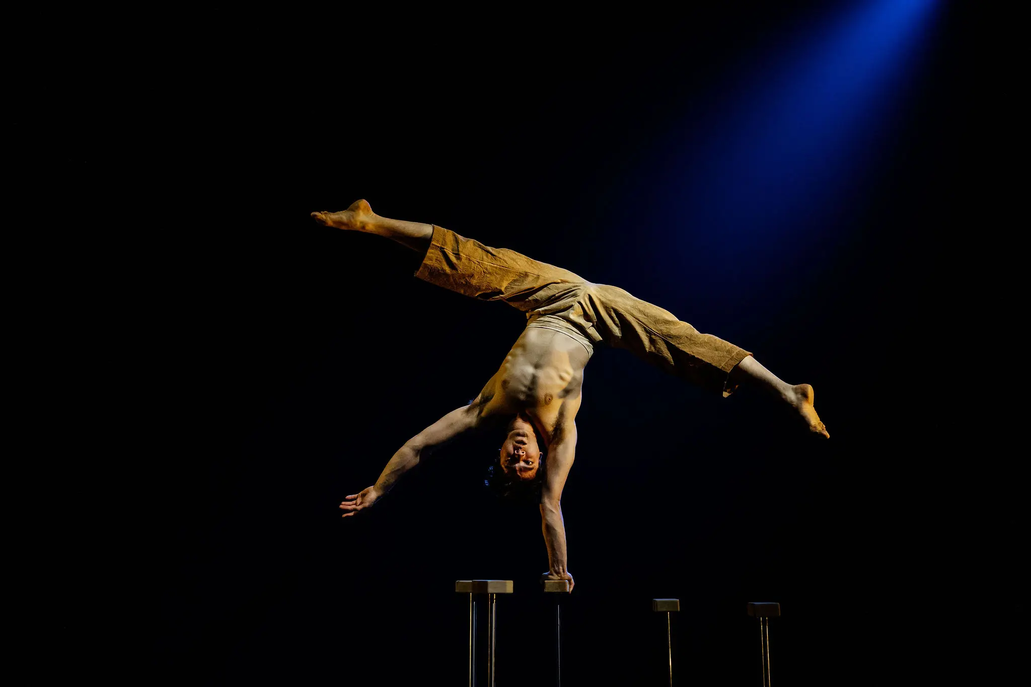 Maël Trân, acrobate torse nu en pantalon marron, effectue un équilibre à une main sur une plate-forme surélevée sous un éclairage de scène spectaculaire, mis en valeur par un projecteur bleu en arrière-plan.