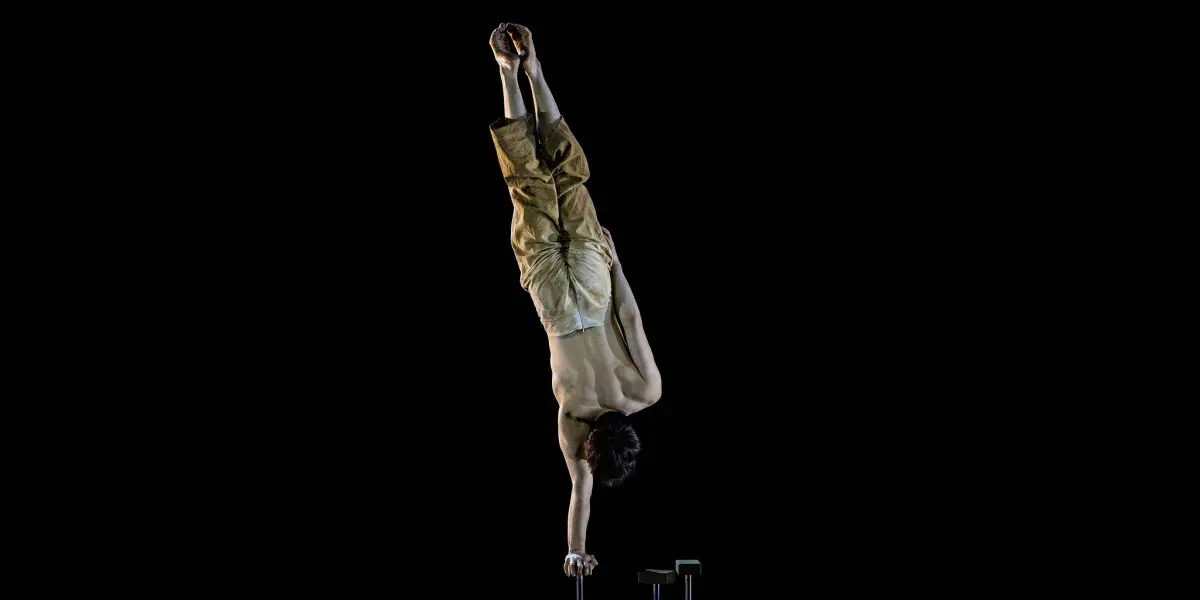 Maël Trân se tient en équilibre sur une main au sommet d'un grand poteau sur un fond sombre, le corps allongé à la verticale et les jambes jointes, vêtu d'un pantalon ample de couleur beige.