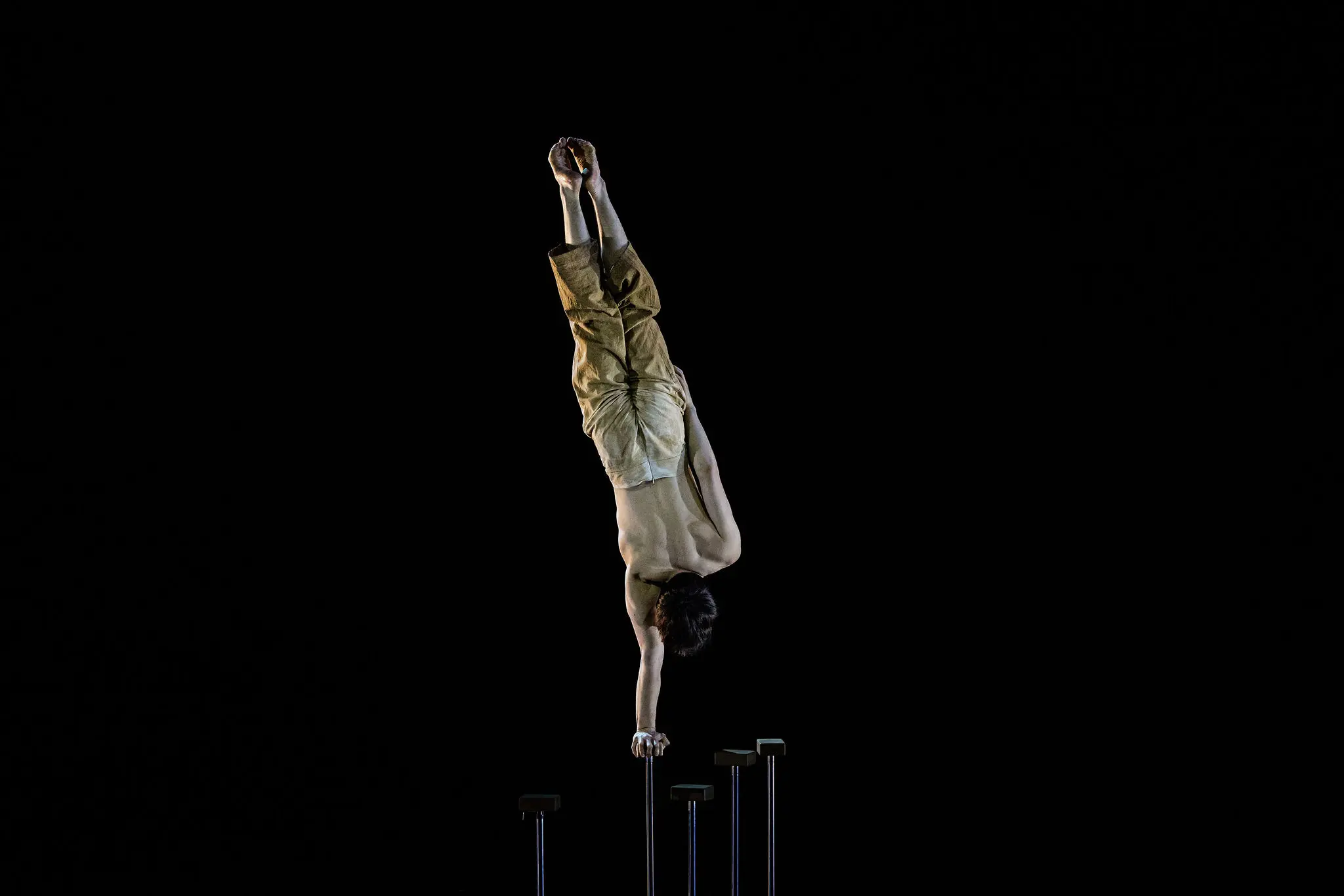 Maël Trân se tient en équilibre sur une main au sommet d'un grand poteau sur un fond sombre, le corps allongé à la verticale et les jambes jointes, vêtu d'un pantalon ample de couleur beige.