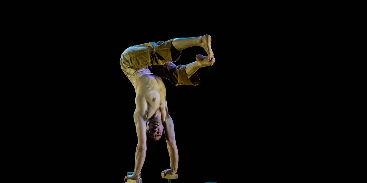 Maël Trân réalise un aplomb torse nu à une main sur des barres parallèles sur fond noir, en équilibre avec les jambes pliées et les pieds croisés en l'air.