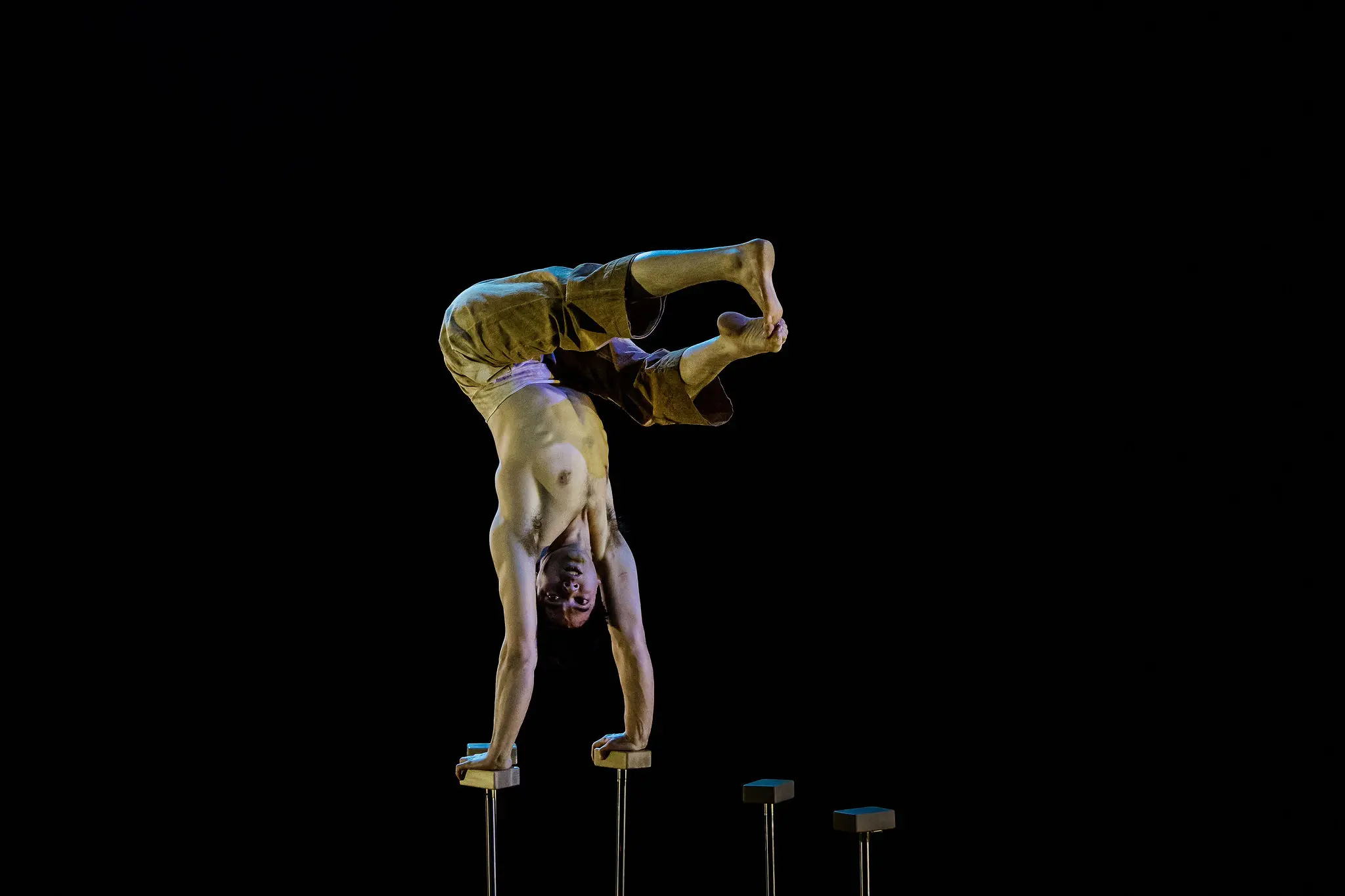 Maël Trân réalise un aplomb torse nu à une main sur des barres parallèles sur fond noir, en équilibre avec les jambes pliées et les pieds croisés en l'air.