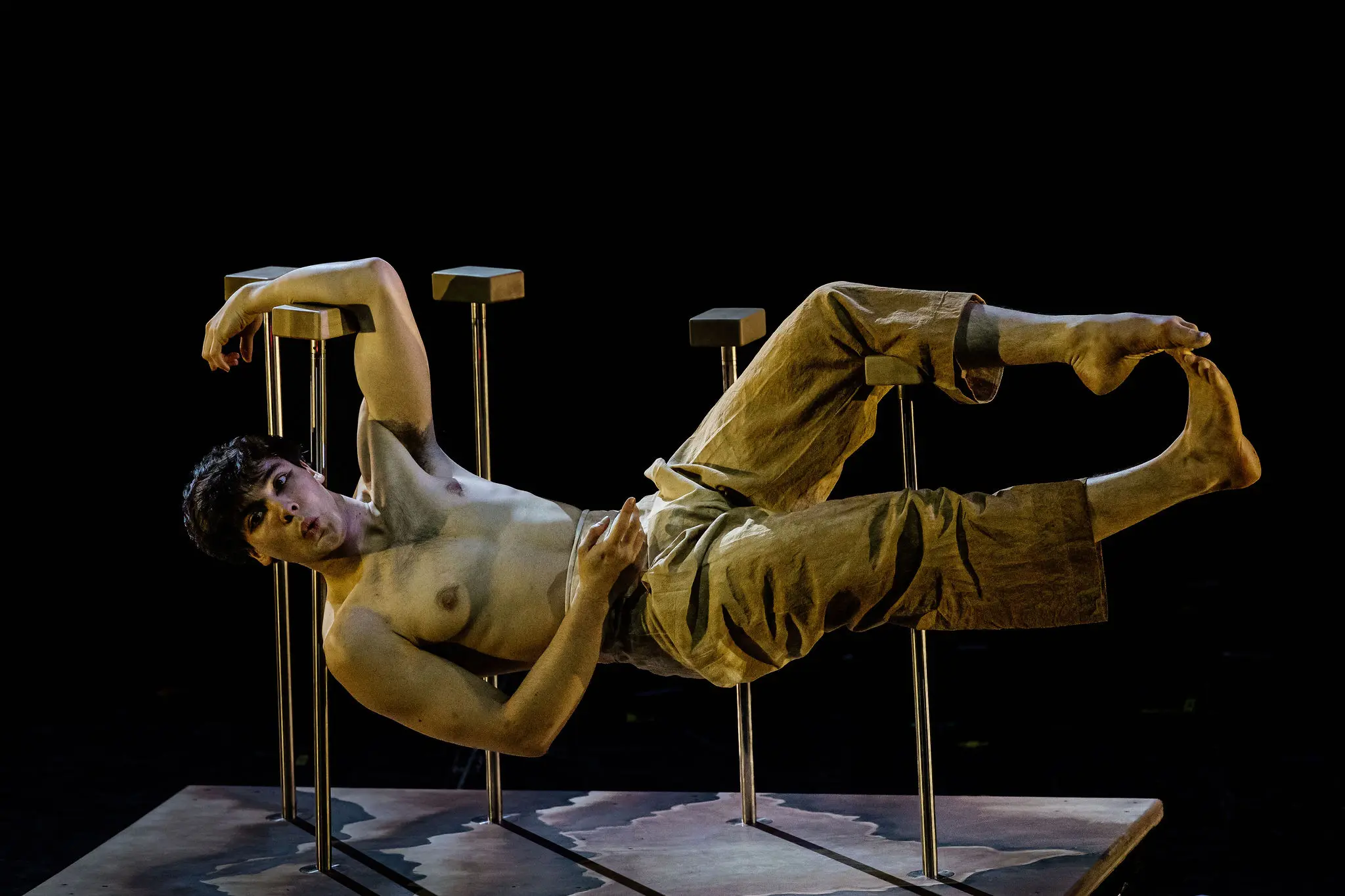 Maël Trân, torse nu et en pantalon ample, se tient en équilibre horizontal sur des tabourets métalliques, soutenu par un bras et une jambe, sur une scène sombre à l'éclairage dramatique.