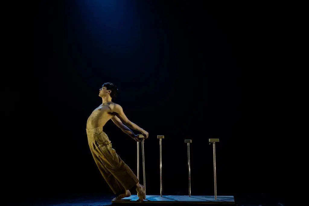 Un artiste torse nu, vêtu d'un pantalon ample, se penche vers l'arrière, saisissant des barres parallèles sur une scène sous les projecteurs -aël Trân attire l'attention, entouré d'obscurité.